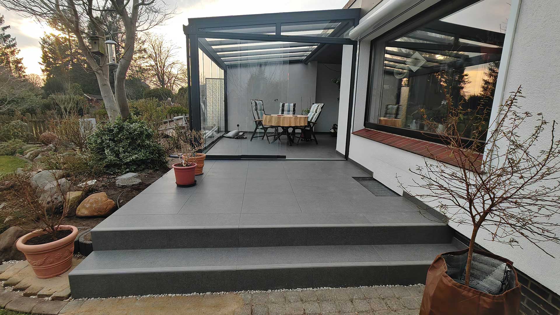 Wintergarten mit grauen Fliesen, Tisch und Stühlen; Pflanzen im Vordergrund; bewölkter Himmel.