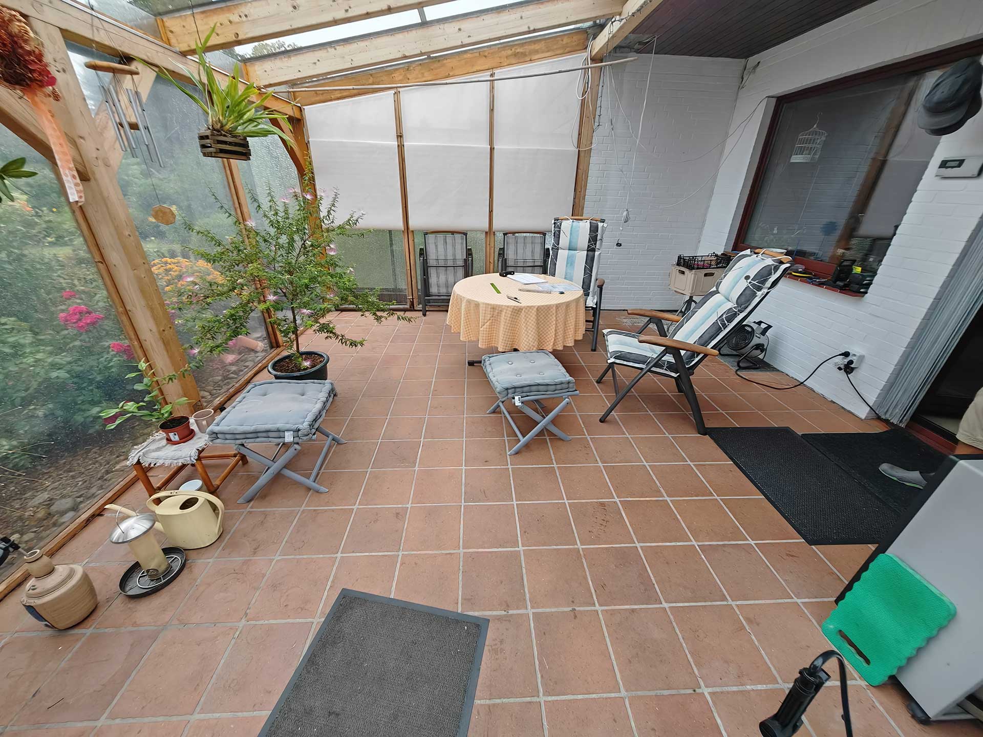 Wintergarten mit Terrakottafliesen, Tisch, Pflanzen, Stühlen und Fenster mit Sicht nach außen.