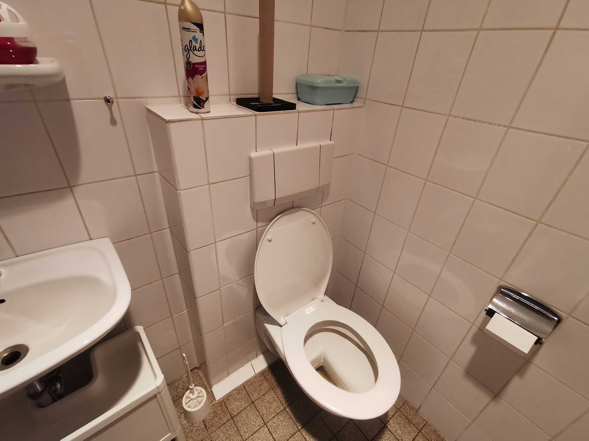 Badezimmer mit Toilette, weiße Fliesen, Duftspray und kleinem Waschbecken rechts im Bild.