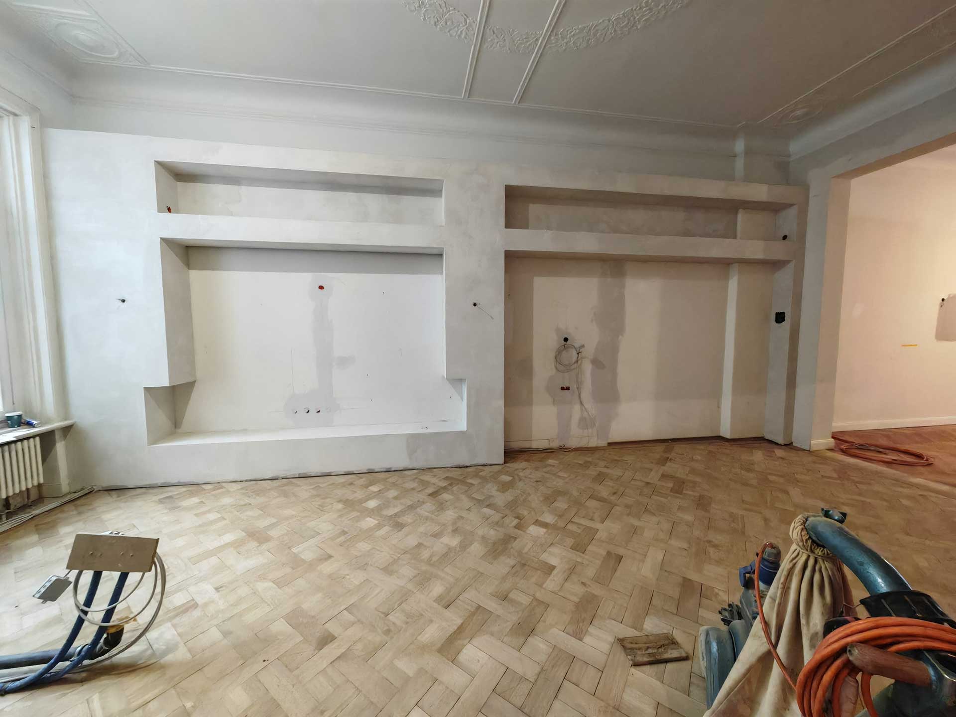 Renoviertes Zimmer mit Parkettboden, Bauwerkzeuge und weiße Wand mit Regaleinschnitten.