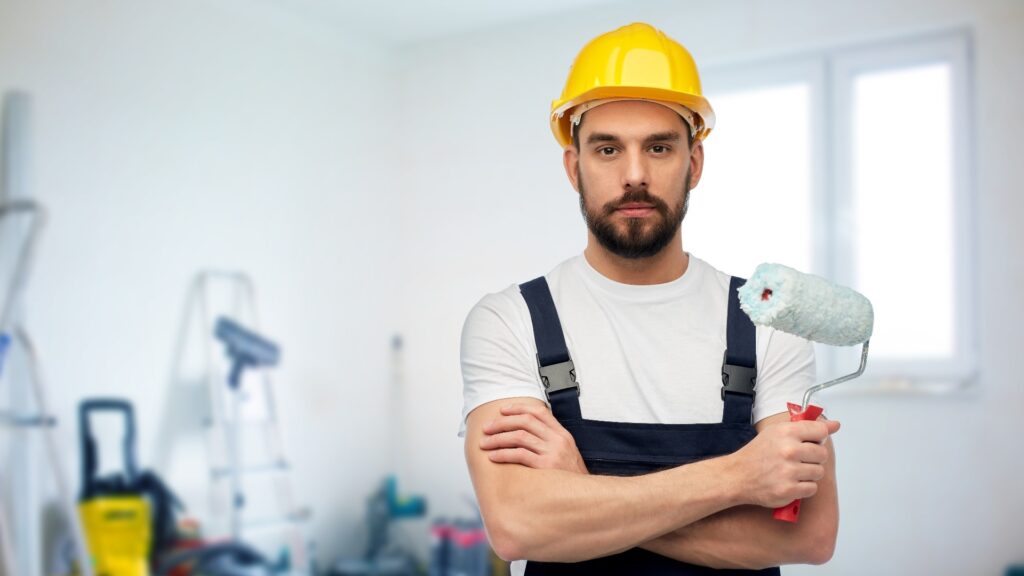 männlicher Arbeiter in gelbem Helm und Overall mit Farbrolle über einem Raum mit Geräten im Hintergrund